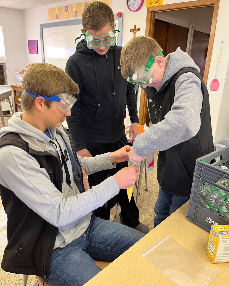 Marek Dvorak holds the bag with a yellow substance inside while Mason Troidl adds chemicals to the mixture. Ezra Sholes looks on, instructing the other two.